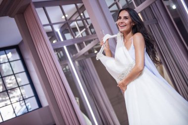 Güzel genç gelin modern düğün salonunun zarif gelinlik seçerken gülümsüyor