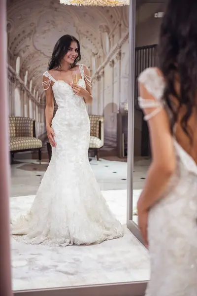 Beautiful young bride is holding a glass of champagne, looking into the mirror and smiling while trying on wedding dress in modern wedding salon