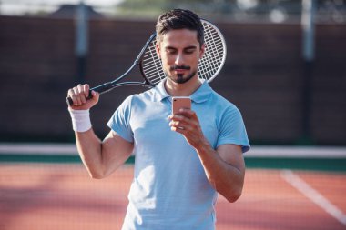 Yakışıklı adam açık havada tenis kortunda Tenis oynarken bir akıllı telefon kullanıyor