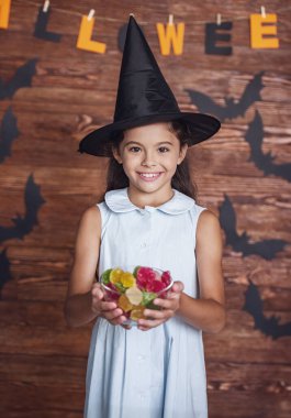 Sevimli küçük kız cadı şapkalı bir kase tatlılar holding, kameraya bakıyor ve Cadılar Bayramı için dekore edilmiş arka plan üzerinde gülümseyen