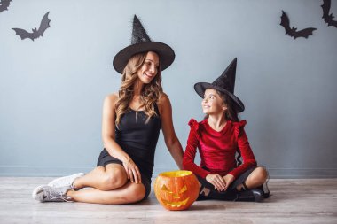 Sevimli küçük kız ve annesi Cadılar Bayramı partisi için ve aksesuarları ile giymiş birbirine bakarak ve yerde otururken gülümseyen