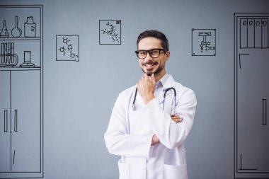 Tıp önlüklü ve gözlüklü yakışıklı doktor kameraya bakıyor ve gülümsüyor, gri arka planda ofis mobilyaları çiziyor.