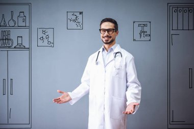 Tıp önlüklü ve gözlüklü yakışıklı doktor kameraya bakıyor ve gülümsüyor, gri arka planda ofis mobilyaları çiziyor.