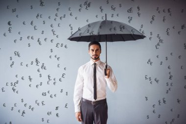 Şemsiyeli yakışıklı işadamı kameraya bakıyor, bankalar yağmur gibi yağıyor, gri arka planda.