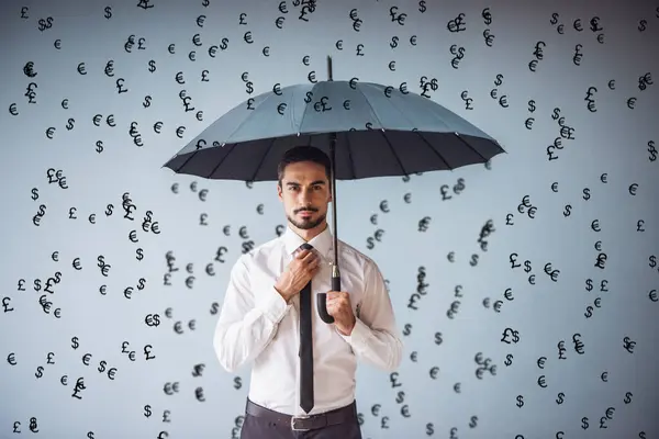 Handsome businessman in classic suit with umbrella is looking at camera, drawn banknotes are falling like rain, on gray background