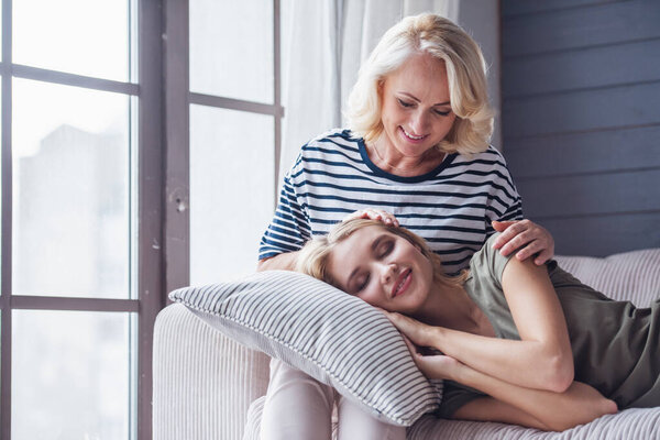 Beautiful senior mom is stroking her adult daughter that lying on her knees, both are smiling while sitting on couch at home