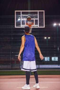 Şık genç basketbolcu geceleri dışarıda oynarken bir topu delikten çekim şapkalı tam uzunlukta portresi