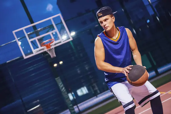 Stylish young basketball player in cap is playing basketball outdoors in the evening