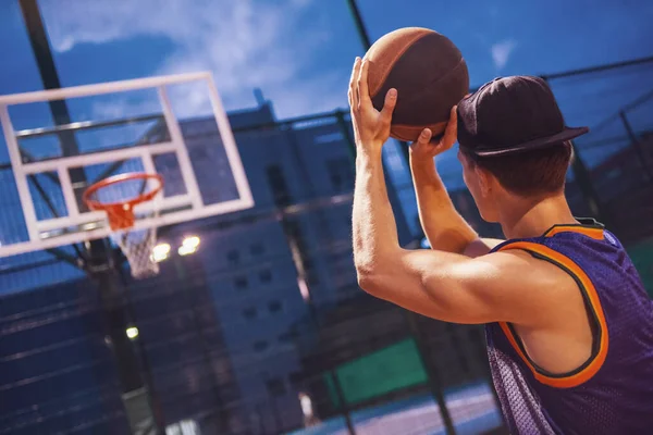 Stylish young basketball player in cap is shooting a ball through a hoop while playing on basketball court outdoors in the evening