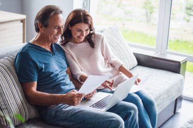 Güzel çift bir dizüstü bilgisayar kullanarak, belgeleri eğitim ve evde kanepede çalışırken gülümseyen