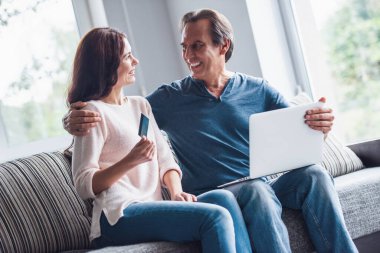 Güzel çift bir dizüstü bilgisayar kullanarak çevrimiçi alışveriş yapıyor ve evde kanepede otururken gülümseyen, kadın bir kredi kartı tutuyor