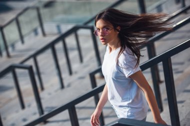 Şık siyah saçlı kız rahat kıyafetler ve gözlük belgili tanımlık merdiven açık havada giderken kameraya bakıyor