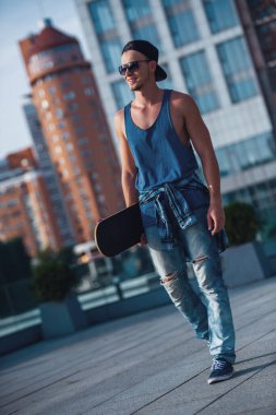 Şık adam rahat kıyafetler, şapka ve gözlük bir kaykay tutarak ve şehirde yürürken gülümseyen