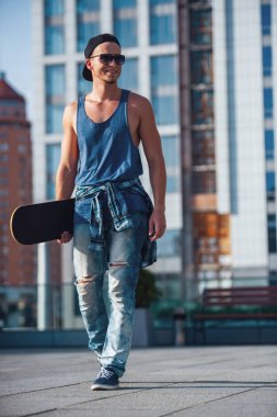 Şık adam rahat kıyafetler, şapka ve gözlük bir kaykay tutarak ve şehirde yürürken gülümseyen