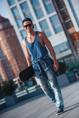 Şık adam rahat kıyafetler, şapka ve gözlük bir kaykay tutarak ve şehirde yürürken gülümseyen