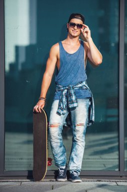 Şık adam rahat kıyafetler, şapka ve gözlük bir kaykay tutarak ve şehirde yürürken gülümseyen