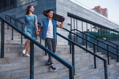 Şık genç çift elele geziyor ve açık havada yürürken gülen, adam bir kaykay tutuyor