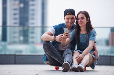 Şık genç çift sarılma, bir akıllı telefon kullanarak ve açık havada kaykay üzerinde otururken gülümseyen