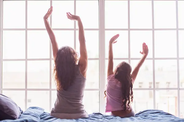 Back view of mom and daughter stretching and yawning while sitting on bed at home