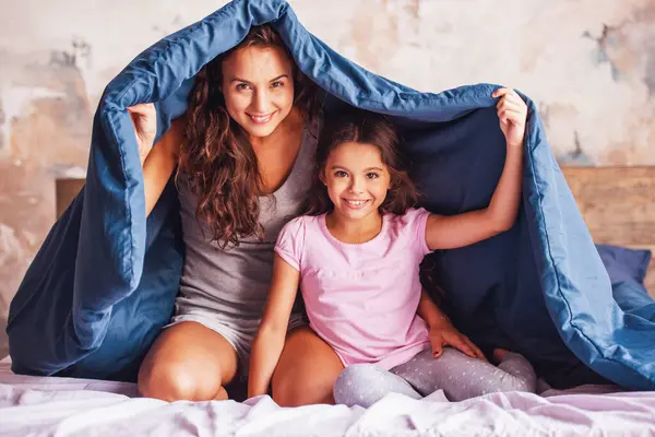 Beautiful mom and daughter are looking at camera and smiling while sitting on bed covered with a duvet