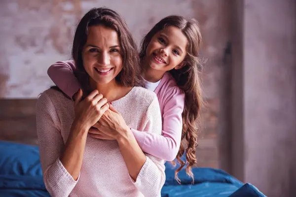 Beautiful mom and daughter are hugging, looking at camera and smiling while sitting on bed at home