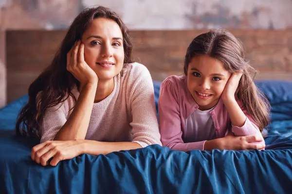Beautiful mom and daughter are looking at camera and smiling while lying on bed at home
