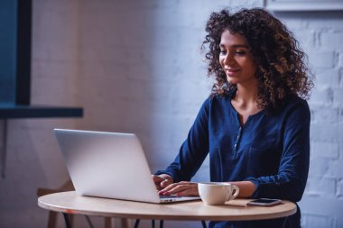 Çekici genç kadın rahat kıyafetler içinde bir dizüstü bilgisayar kullanarak ve kafede istirahat ederken gülümseyen