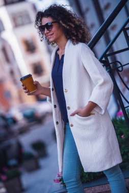 Beyaz önlük ve Güneş gözlükleri çekici genç kadın kahve içme ve şehirde yürürken gülümseyen