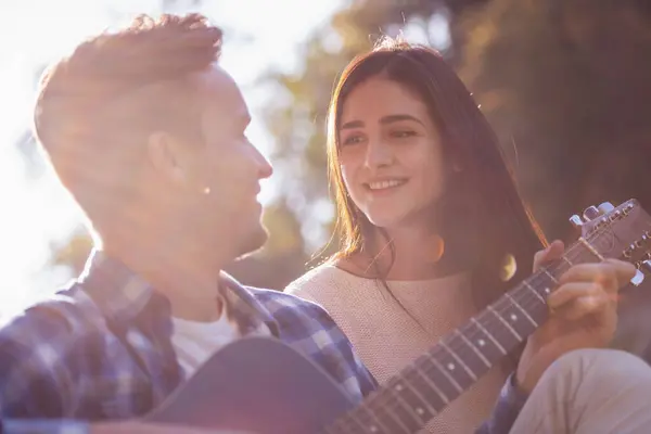 Beautiful young couple is singing and smiling while resting on a plaid in the park, guy is playing guitar