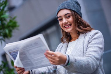 Güzel kız ceket ve kap bir gazete okuyor ve açık havada dinlenme süre gülümseyen