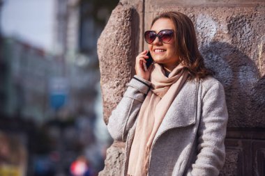 Ceket ve güneş gözlüklü güzel kız cep telefonda konuşurken ve şehirde yürürken gülümseyen