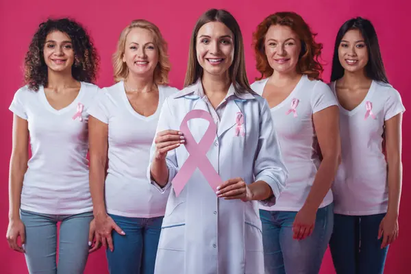 妇女反对乳腺癌 不同年龄和民族的美丽的妇女在他们的胸口微笑的粉红色丝带 医生拿着粉红色的纸丝带 在红色背景 — 图库照片