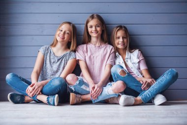 Rahat kıyafetler içinde üç çekici genç kız kameraya bakarak ve gri duvar zemin katta otururken gülümseyen