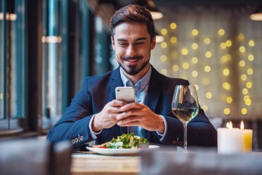 Takım elbiseli yakışıklı işadamı bir akıllı telefon kullanarak ve restoranda otururken gülümseyen