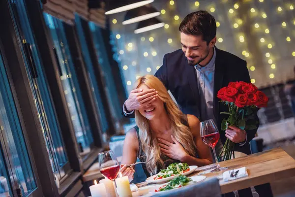 Handsome elegant man is holding roses and covering his girlfriend\'s eyes while making a surprise in restaurant, both are smiling