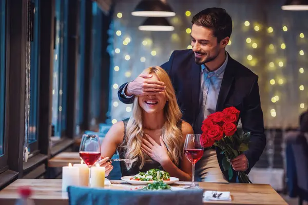 英俊优雅的男子捧着玫瑰花 遮住女友的眼睛 在餐厅里做着惊喜 两人都在微笑 — 图库照片