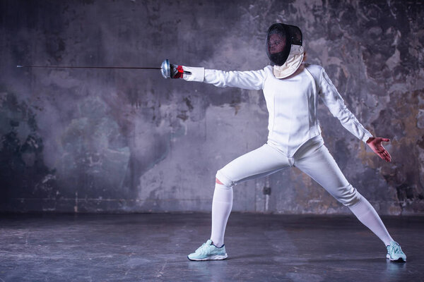 Женщина-фехтовальщик в защитной одежде использует оружие, практикуясь на темно-сером фоне

