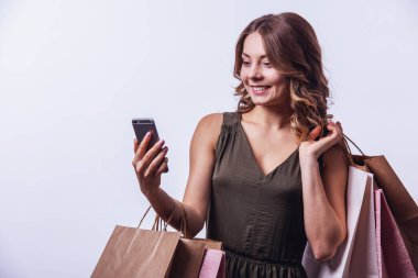 Elbiseli çekici genç kadın alışveriş torbaları tutarak, bir smartphone kullanarak ve, ışık arka plan üzerinde gülümseyen