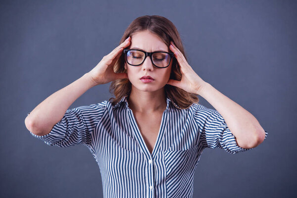 Привлекательная уставшая молодая женщина в очках трогает голову на сером фоне.
