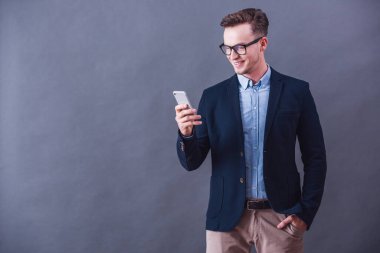 Takım elbise ve gözlük yakışıklı genç işadamı bir akıllı telefon kullanarak ve gri arka plan üzerinde gülümseyen