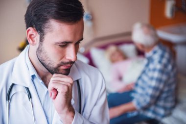 Yaşlı adam ön planda yakışıklı doktor hastane koğuşunda yatakta yatan karısını ziyareti yapıyor