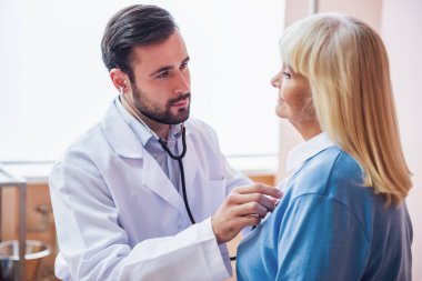 Yakışıklı Doktor bir stetoskop kullanarak yaşlı bir kadın kalp atışına dinleme