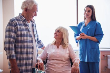 Güzel yaşlı kadın kocasına hastane koğuşunda tekerlekli sandalyede otururken konuşuyor, Hemşire yakınındaki duruyor, tüm gülümsüyor musun