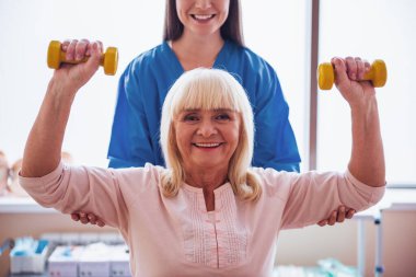 Güzel yaşlı kadın çalışmaları ile halter, kameraya bakarak ve, gülümseyen hastane koğuşunda yapıyor. Çekici hemşire ona yardım ediyor