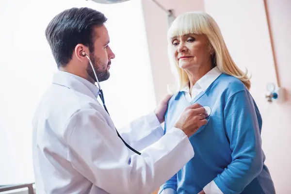 Красивый Доктор Слушает Сердцебиение Пожилой Женщины Используя Стетоскоп Стоковое Изображение