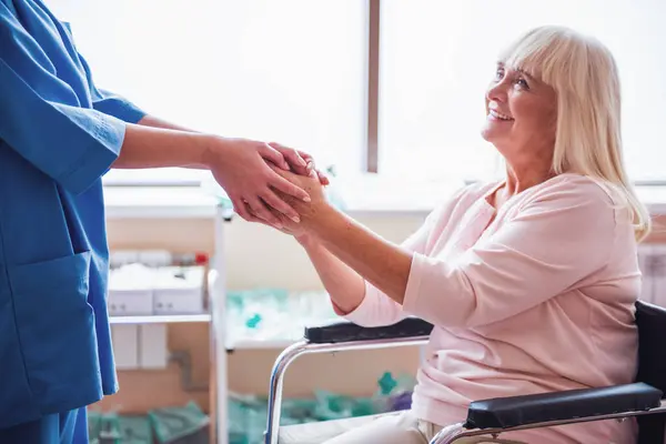 美丽的老太太握着医生的手 微笑着坐在医院病房的轮椅上 免版税图库图片