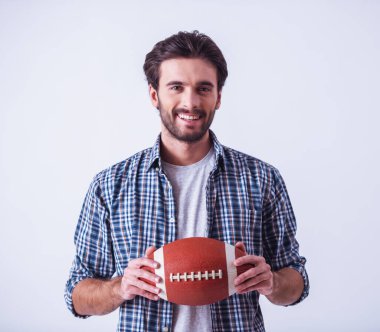 Rahat kıyafetler içinde yakışıklı sakallı adam kameraya bakıyor ve gülümsüyor, açık renkli bir top tutuyor