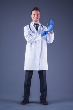 Yakışıklı genç doktor eldiven koyarak, kameraya bakıyor ve gülümsüyor, gri arka plan üzerinde tıbbi ceket tam boy görüntü