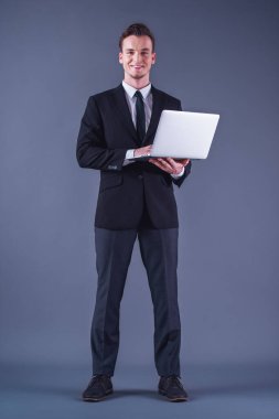 Yakışıklı genç iş adamı bir dizüstü bilgisayar kullanarak, kameraya bakıyor ve gülümsüyor, gri arka plan üzerinde elbise tam boy görüntü