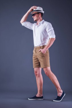 Tam uzunlukta, arka görünümü görüntü yakışıklı genç adamın şapka koyarak ve gri arka plan üzerinde yürürken gülümseyen gömlek, şort ve güneş gözlüklü